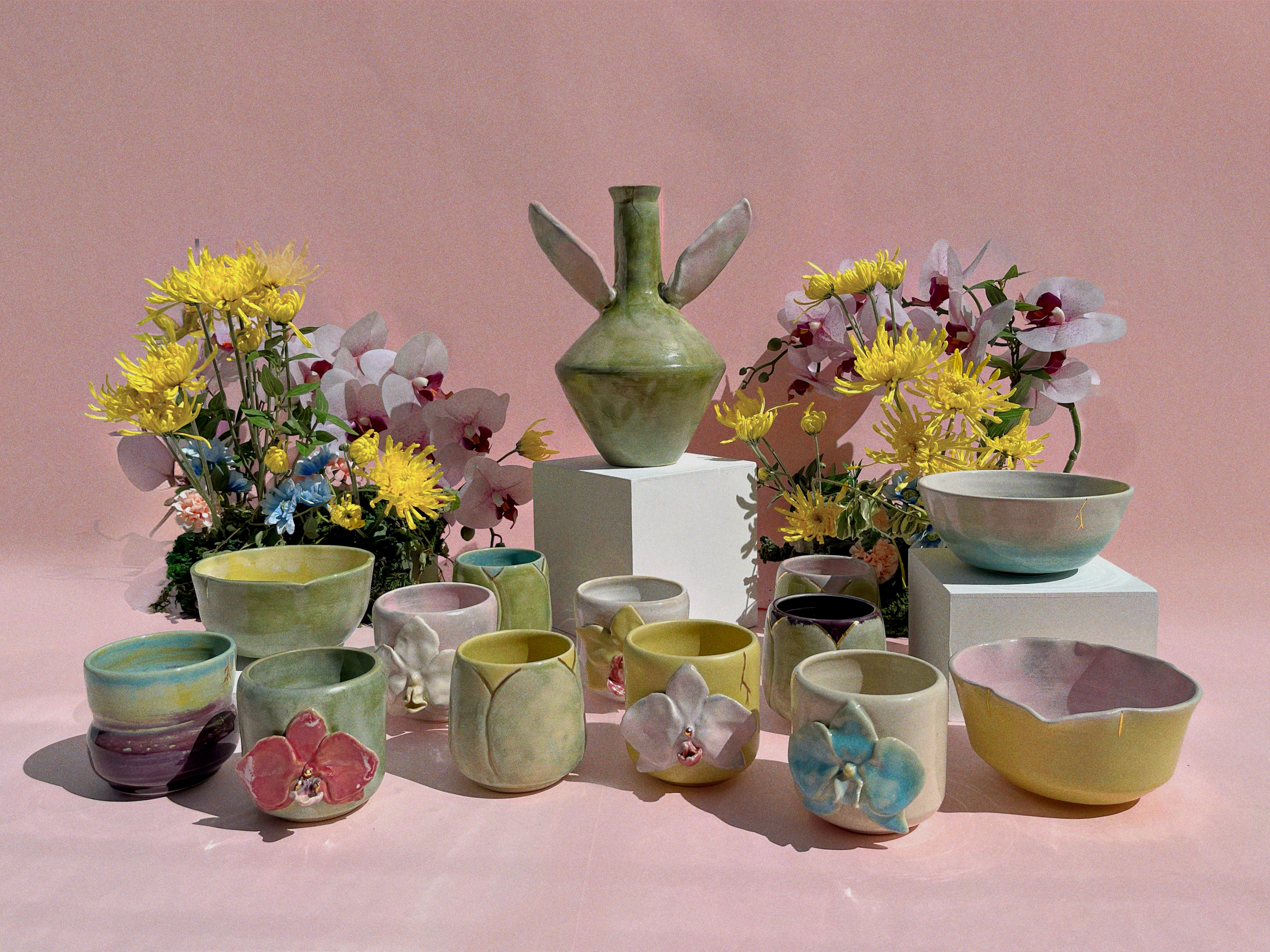 A collection of handmade ceramic tableware, including bowls, cups, and a winged vase, displayed on white pedestals with decorative flower arrangements against a solid pink background.