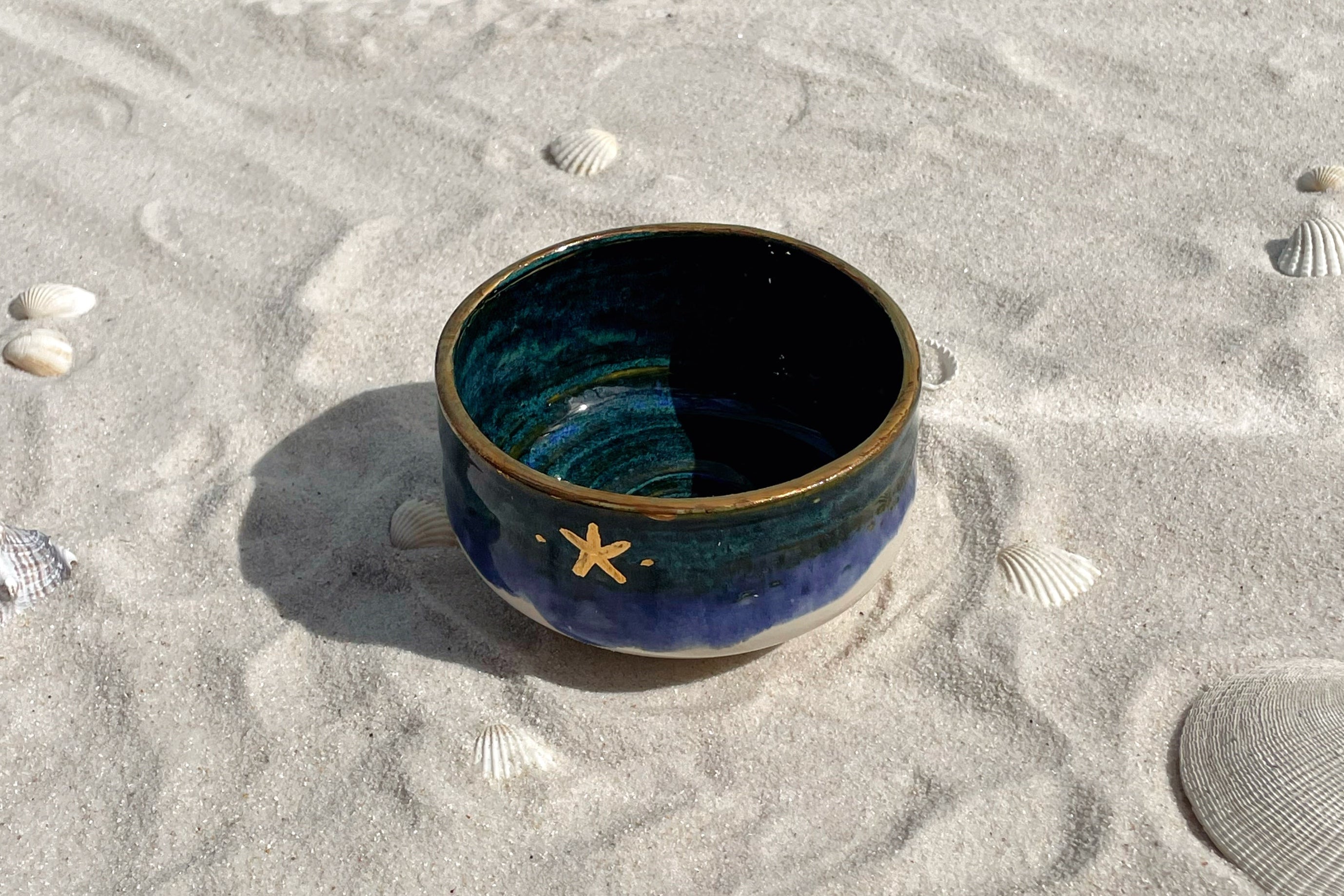 Blue ceramic bowl with a starfish design on a sandy surface with seashells.